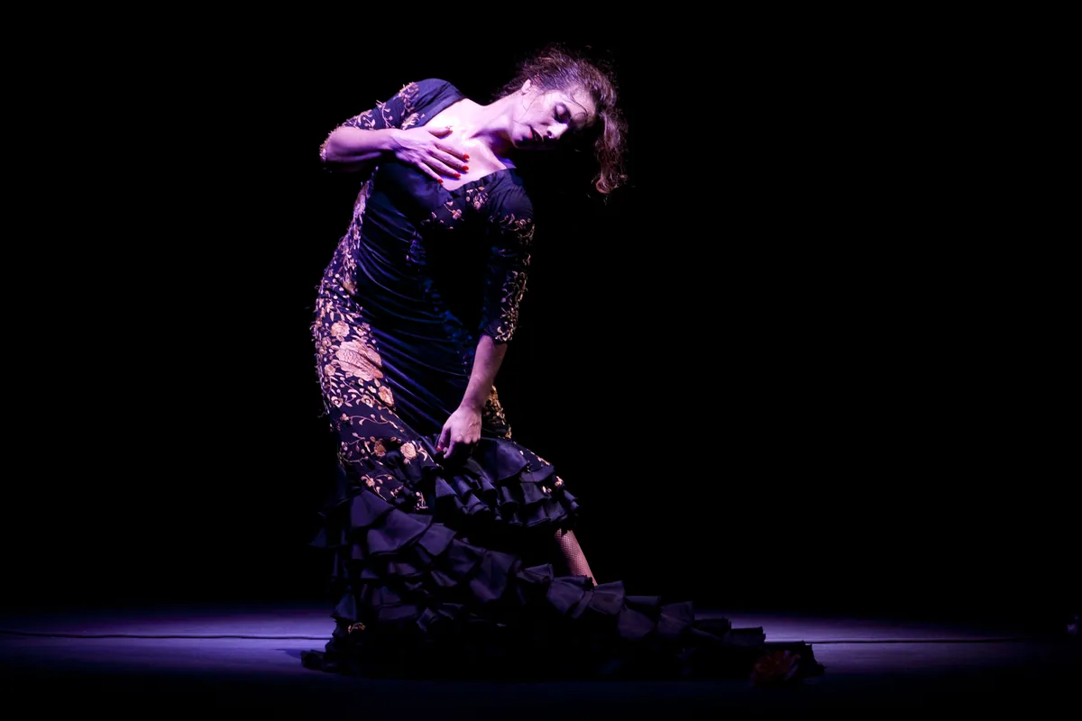 A flamenco dancer in a dark embroidered dress bending from the waist with one hand on her chest, alone in a spotlight on a black stage
