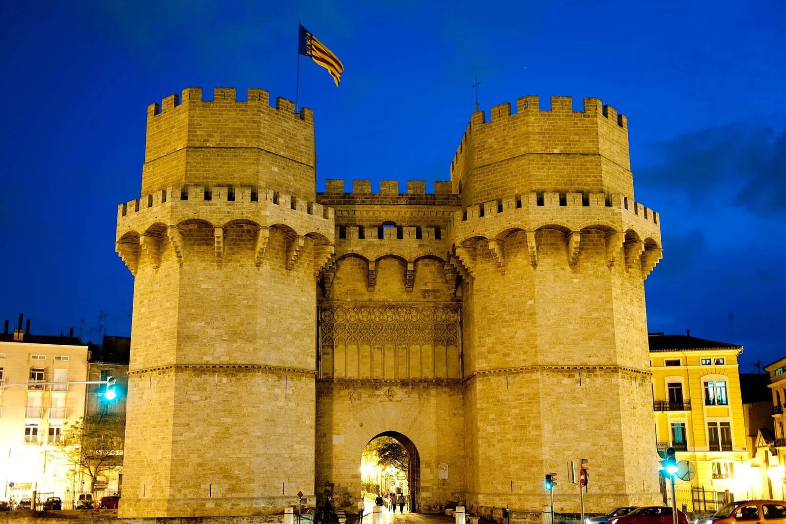 The Torres de Serranos at dusk, with a Valencian flag on the left tower and yellow-lit stone under a deep blue sky